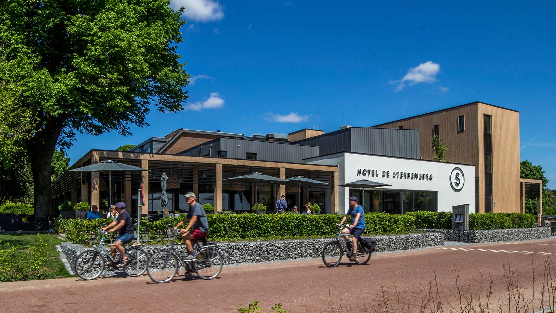 Gebouw HOTEL DE STERRENBERG toont houten gevel en terras; fietsers rijden voorbij in groene omgeving bij De Hoge Veluwe.